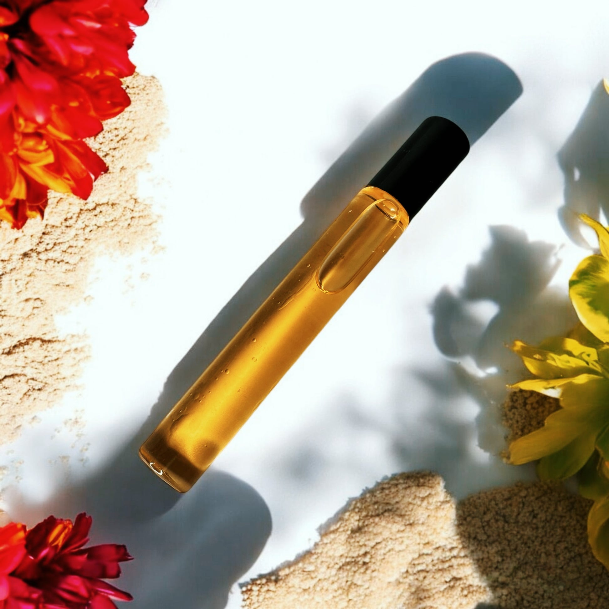 A 10ml roller ball perfume bottle with a golden hue, displayed on a sandy surface with colorful flowers around it.